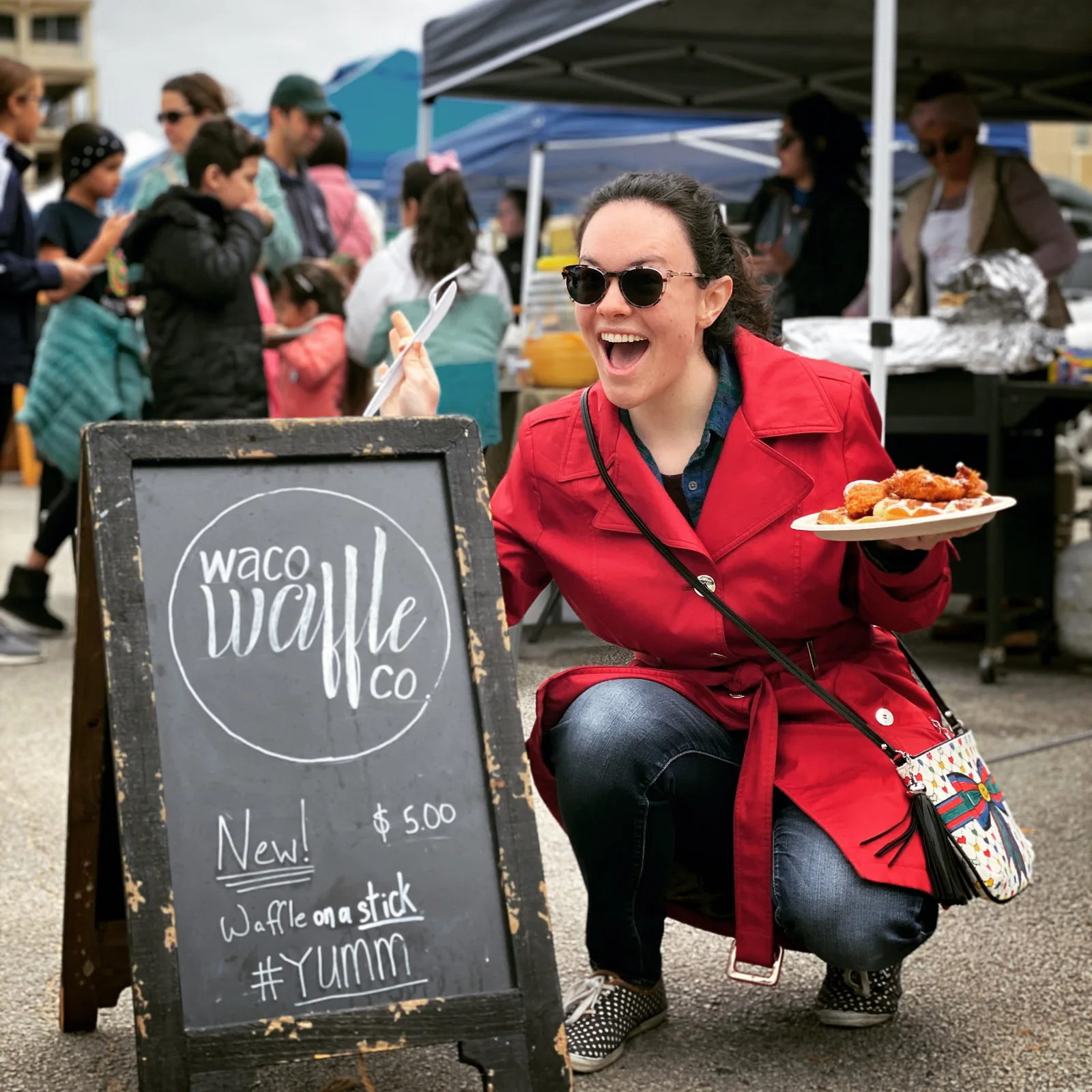 Excited customer in a red coat crouching beside the Waco Waffle Co. chalkboard sign, holding a plate of chicken and waffles