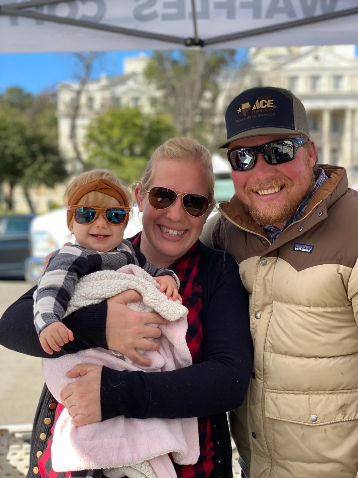 Seth and his wife smiling under the Waco Waffle Co. tent, holding their baby wearing sunglasses