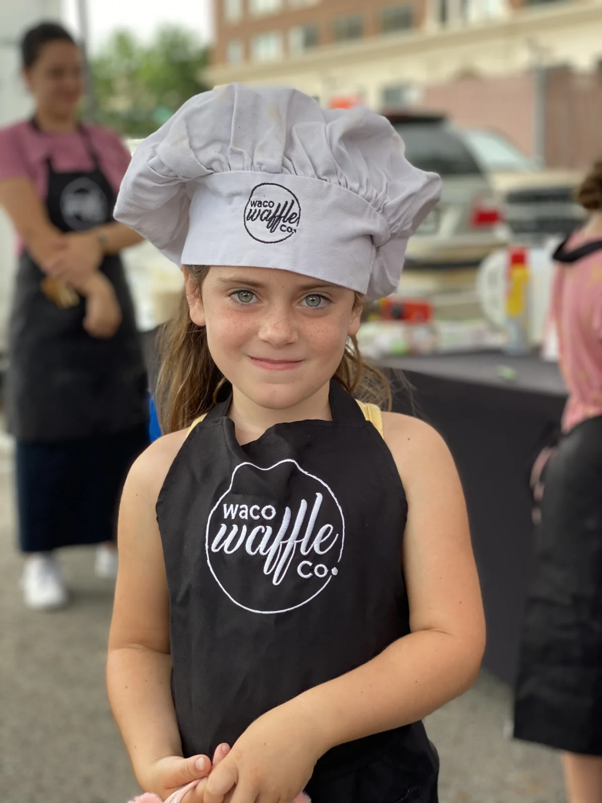 Young girl smiling in a Waco Waffle Co. chef hat and black apron at the market